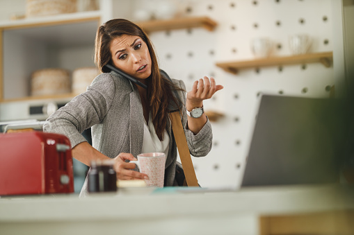 Shot of a multi-tasking young business woman talking on her smartphone and drinking coffee in her kitchen while getting ready to go to work.