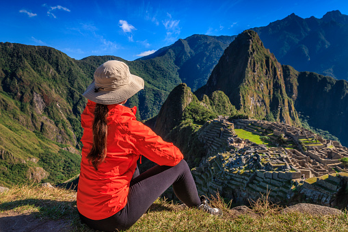 Young female tourist, wearing an orange jacket, is looking at Machu Picchu. Machu Picchu (Quechua: Machu Picchu) – \