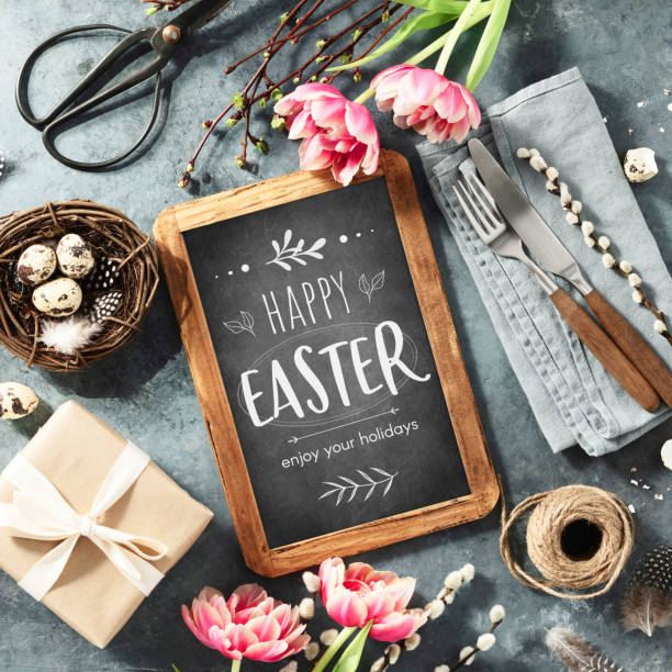 Easter table setting with Happy Easter text on vintage chalk board, spring flowers and cutlery on dark blue background top view flat lay stock photo