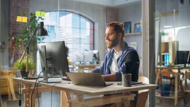 istock Handsome Long-Haired Bearded Manager Sitting at a Desk in Creative Office. Young Stylish Man Using Laptop Computer with Another Monitor in Marketing Agency. Colleagues Working in the Background. 1479753797