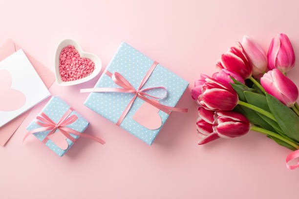 concetto di festa della mamma. foto in alto di scatole regalo blu con fiocchi a nastro bouquet di tulipani rosa piattino a forma di cuore con spruzzate e busta con cartolina su sfondo rosa pastello isolato - grace tulip flower pink foto e immagini stock