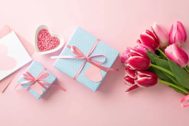 Photo of Mother's Day concept. Top view photo of blue gift boxes with ribbon bows bouquet of pink tulips heart shaped saucer with sprinkles and envelope with postcard on isolated pastel pink background