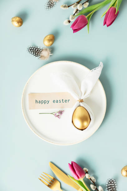 istock Stylish Easter flat lay with golden egg in easter bunny napkin on white plate, golden quail eggs, feathers, cutlery and spring flowers on blue background. Minimalist modern Easter background 1479761358