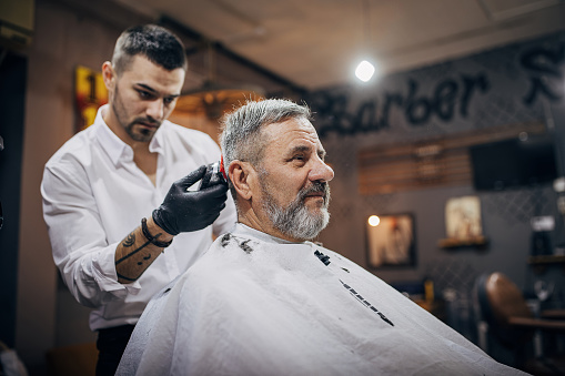 Two people, barber cutting senior man's hair with hair clipper in barber shop.