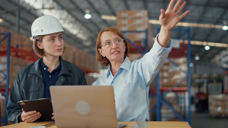 Young caucasian woman worker and mature female manager checks stock inventory with tablet and laptop and discuss talk together in retail warehouse. Logistic industry business.