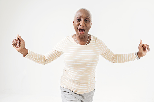 Positive senior female in casual clothes smiling and holding arms up while standing on white background