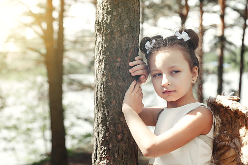 Pensive little girl in angel image with gold wings posing by tree in summer forest, looking away. Thoughtful young lady in sunshine woodland. Environmental protection concept. Copy ad text space