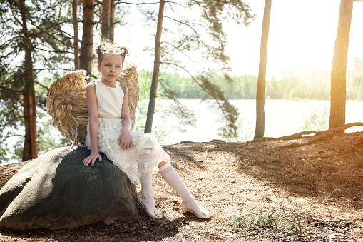 Pensive little girl in white dress with angel gold wings sitting on rock in summer forest, looking away. Thoughtful young lady in sunshine woodland. Environmental protection concept. Copy text space