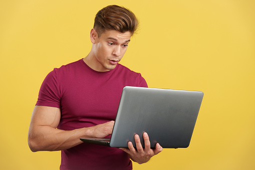Surprised strong man standing using a laptop in studio with yellow background