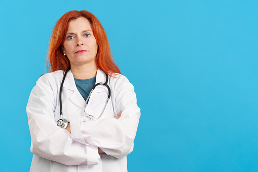 Serious mature redheaded female doctor standing with arms crossed in studio with blue background