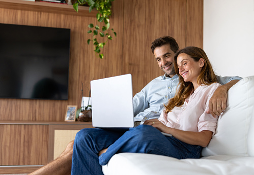 Happy Brazilian couple relaxing at home watching movies online on their laptop and smiling