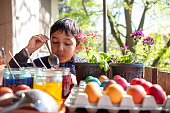 A young woman paints eggs for Easter in a cottage in the forest. A small child plays next to her.