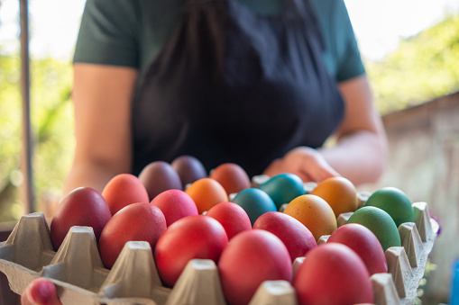 A young woman in a green blouse with short sleeves and a black apron holds a carton package container with eggs painted for Easter.