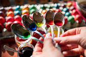 A woman holds spoons of ralsic egg dyes over cardboard containers filled with dyed Easter eggs.