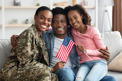 Beautiful black family celebrating reunion at home, happy father and daughter greeting their mom soldier, sitting on couch, embracing and smiling at camera, holding flag of the US
