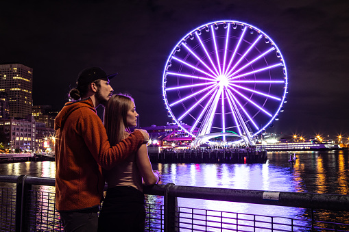 Man and Woman Couple Admiring Seattle Washington at night, The Seattle Great Wheel Lit Up Reflecting on Pacific Ocean, Pier 69 Harbor, Long Exposure, City Lights in the Background