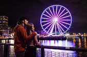 Man and Woman Couple on Vacation Admiring Seattle Washington at night The Seattle Great Wheel Lit Up Reflecting on Pacific Ocean Pier 69 Harbor Long Exposure City Lights in the Background