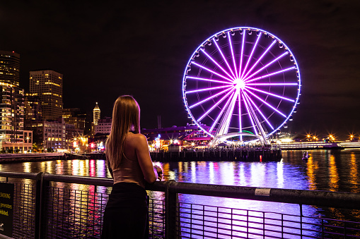 Woman Standing Alone Looking at Seattle Washington at Night, The Great Seattle Wheel Lit Up, Reflecting on Pier 69 Harbor in the Pacific Ocean, Long Exposure Photo with City Lights in the Background