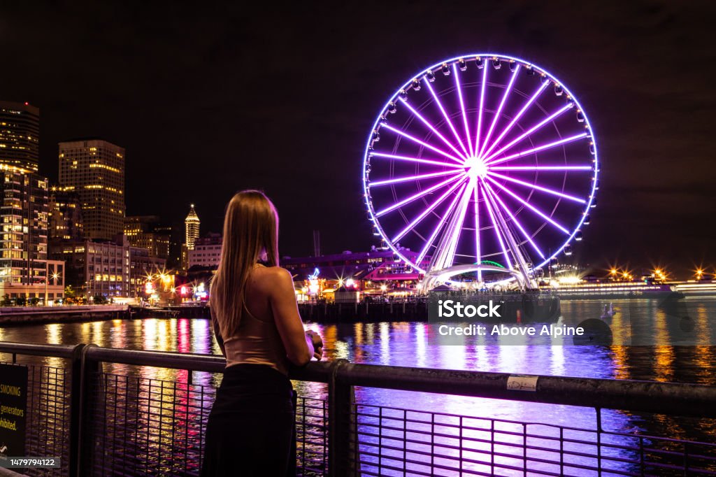 Woman Standing Alone Looking at Seattle Washington at Night The Great Seattle Wheel Lit Up Reflecting on Pier 69 Harbor in the Pacific Ocean Long Exposure Photo with City Lights in the Background Woman Standing Alone Looking at Seattle Washington at Night, The Great Seattle Wheel Lit Up, Reflecting on Pier 69 Harbor in the Pacific Ocean, Long Exposure Photo with City Lights in the Background Seattle Great Wheel Stock Photo