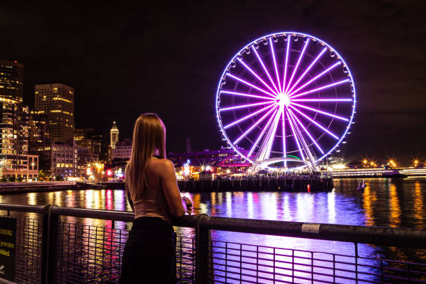 Woman Standing Alone Looking at Seattle Washington at Night The Great Seattle Wheel Lit Up Reflecting on Pier 69 Harbor in the Pacific Ocean Long Exposure Photo with City Lights in the Background Woman Standing Alone Looking at Seattle Washington at Night, The Great Seattle Wheel Lit Up, Reflecting on Pier 69 Harbor in the Pacific Ocean, Long Exposure Photo with City Lights in the Background seattle ferris wheel stock pictures, royalty-free photos & images