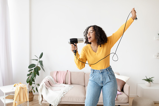 Happy young black lady with hair dryer as microphone singing and dancing at home, copy space. Beautiful African American woman pretending to be rockstar, having fun indoors