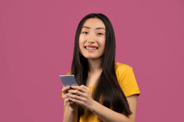 istock Cool gadget and app. Happy korean lady using smartphone and smiling at camera, standing on violet studio background 1479794424