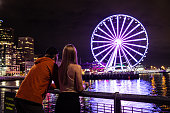 Man and Woman Couple on Vacation Looking at Seattle Washington Skyline at night Long exposure The Seattle Great Wheel Reflecting on Pacific Ocean Pier 69 City Lights in backgroung