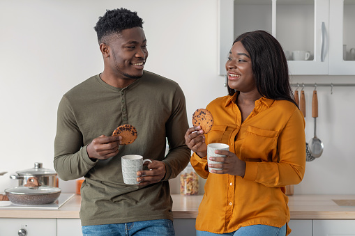 Portrait Of Happy Young Black Couple Drinking Coffee And Eating Cookies In Kitchen, Romantic African American Spouses Enjoying Homemade Snacks And Hot Drinks And Smiling To Each Other, Free Space