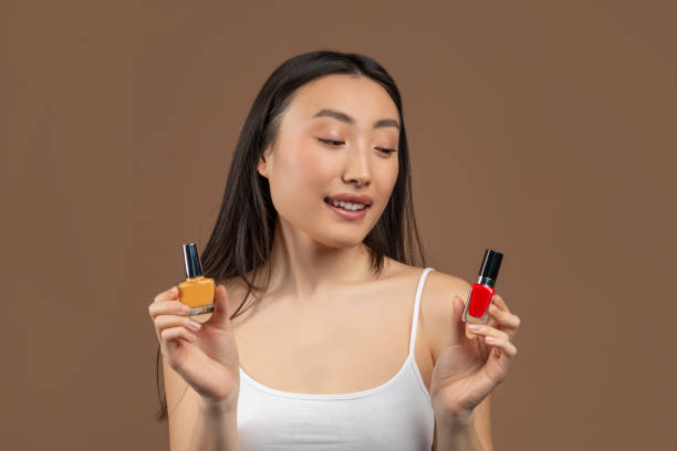 retrato de una joven asiática dudando sobre elegir esmalte de uñas, posando sobre un fondo de estudio marrón - hesitating fotografías e imágenes de stock
