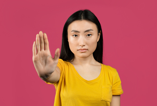 Young japanese lady gesturing STOP, saying NO, showing rejection gesture, expressing her negative attitude, posing on pink studio background
