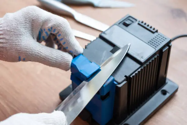 Sharpening a knife on an electric sharpener at home. The man's hand drives the knife blade between the blue sharpeners, dust flies on the machine.