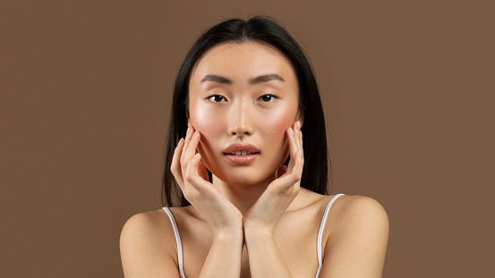Flawless beauty. Portraif of asian lady touching her soft smooth skin on cheeks, looking at camera while standing on brown studio background, panorama
