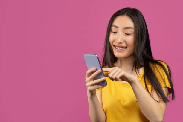 istock Cheerful asian lady using smartphone for messaging with friends, texting on cellphone, violet background, free space 1479797161