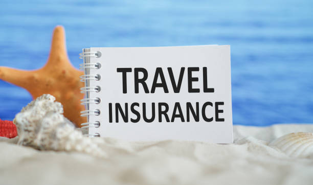 Travel insurance text on notepad with seashells on tropical white sand beach against blue sea - leisure and business concept