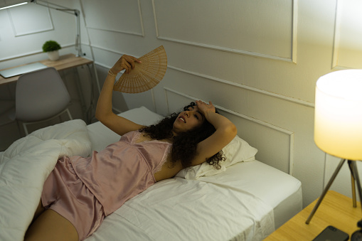 Upset young woman in pajamas feeling hot and using a fan during the summer in bed and having sleeping problems