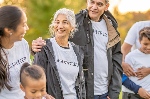 A small group of men, women and children work together outside as they volunteer in their community.  They are each dressed casually and are wearing matching volunteer t-shirts.
