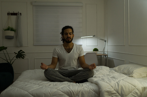 Relaxed hispanic man in pajamas listening to relaxing music with headphones while doing a meditation in bed before sleeping