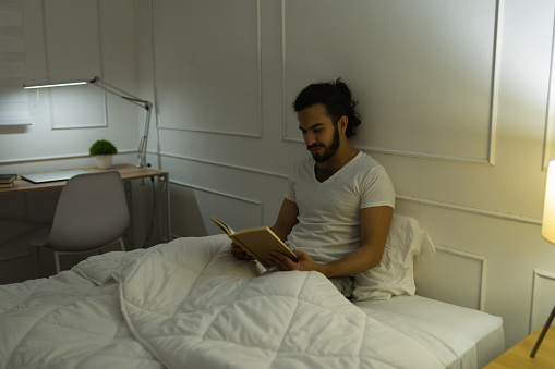 Attractive relaxed young man lying in bed in pajamas while reading an interesting book before sleeping at night
