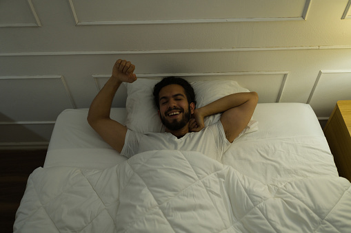 Happy attractive young man smiling and feeling relaxed and well rested after waking up and stretching in bed