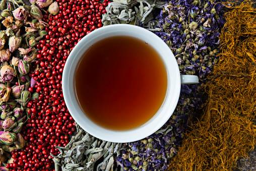 Directly above view of a tea cup with herbal tea over a background made of a variety of herbal teas and tea ingredients like Red pepper, Chamomile tea, Sage tea, Linden tea, Fennel tea, Melissa tea, Rose tea,  Rosemary Tea , Corn silk tea , dried orange slices and rosemary on wooden background.