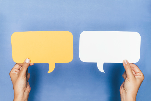 Two hands are holding two speech bubbles which are white and yellow in front of blue background.