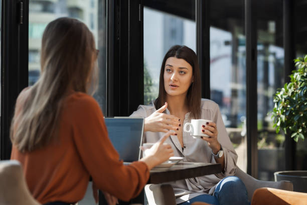 Two young business women in a cafe having one on one meeting. Friends after work talking gossiping and having coffee at a window table on a sunny day.. stock photo