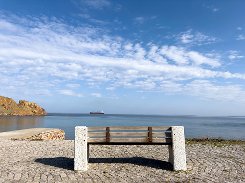 A detailed capture unveils the rustic beauty of a half-buried, weathered barrel in the sandy embrace of Coughlin Park, Winthrop, just outside Boston, Massachusetts, USA. Bathed in sunlight, the scene unfolds against a backdrop of shimmering water and sailboats, weaving a tale of coastal allure and time's gentle touch.