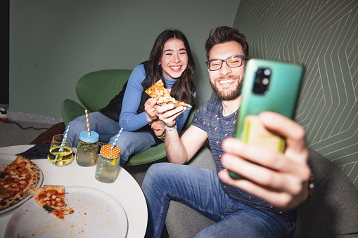 An attractive happy young adult Caucasian male taking a selfie with his female best friend at the bar. They are eating a delicious pizza while taking a selfie. The friends are smiling and enjoying their time together hanging out. They are taking a photo to commemorate the fun times they have together.