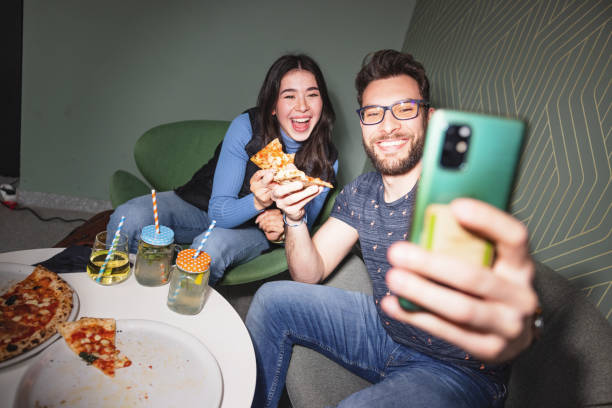 счастливый привлекательный молодой взрослый кавказский мужчина делает селфи со своей подругой в баре, наслаждаясь пиццей - mobile phone flash стоковые фото и изображения