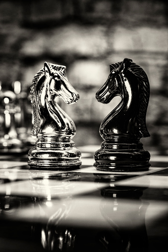 Two opposing knights positioned in front of each other on a fully set up chess board, shot in studio against a brick background suggesting a metaphor for confrontation or battle.