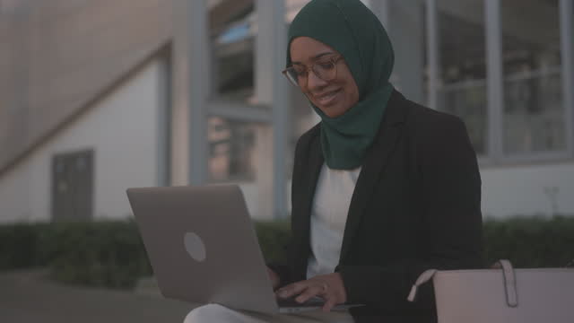 Smiling muslim businesswoman sitting on a bench outdoors and using a laptop