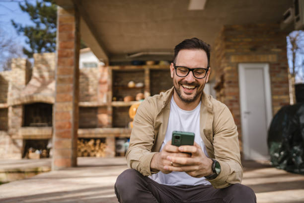 um homem adulto na casa da frente no dia usar chamada de vídeo do telefone inteligente ou sms - mid adult men smart casual white happiness - fotografias e filmes do acervo