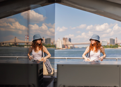 In New York City, a young woman is enjoying a ferry trip on the East River in summer. Woman's reflection is seen in the glass door.
