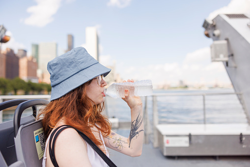 Young woman is riding ferry on East River in New York City and drinking water on a hot, summer day.